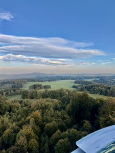 Blick aus großer Höhe auf eine weitläufige, hügelige Waldlandschaft unter blauem Himmel mit vereinzelten Wolken.