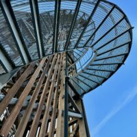 Eine Aufnahme aus tiefem Blickwinkel einer runden Metallaussichtsplattform auf der Spitze eines hohen Holzturms vor einem strahlend blauen Himmel.
