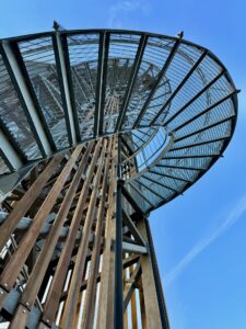 Eine Aufnahme aus tiefem Blickwinkel einer runden Metallaussichtsplattform auf der Spitze eines hohen Holzturms vor einem strahlend blauen Himmel.